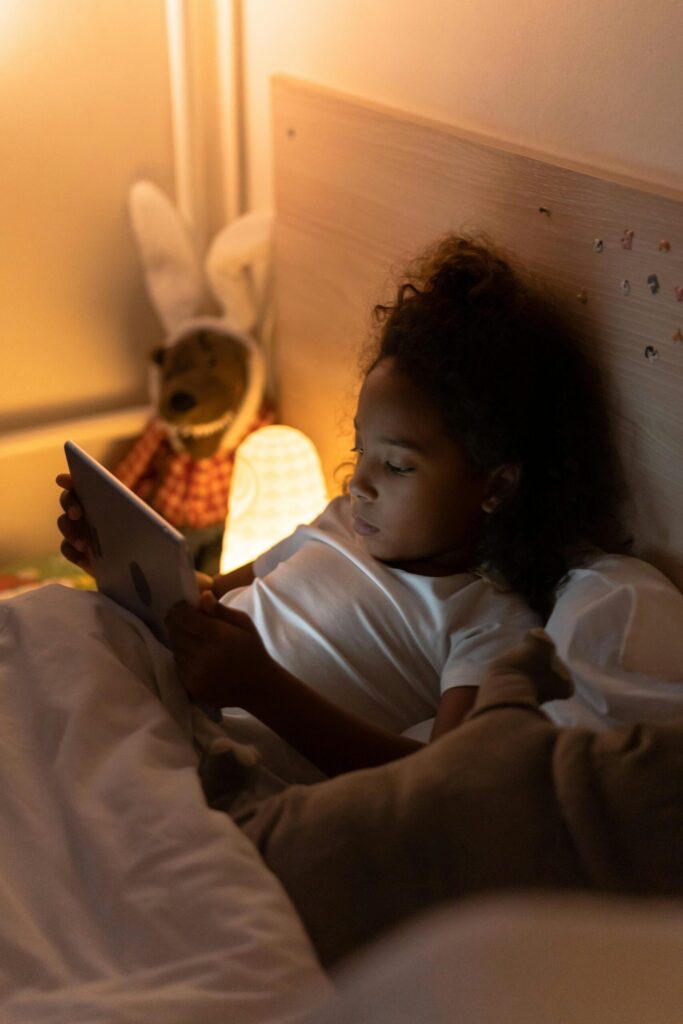 Child enjoying a tablet in a cozy bedroom ambiance with soft lighting and plush toys.
